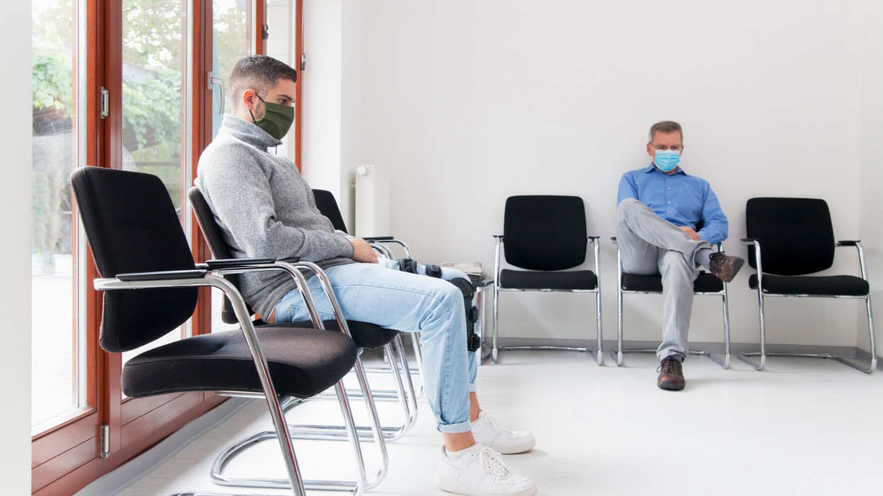 Young and mature man with face masks sitting in a waiting room of a hospital or office - focus on the young man