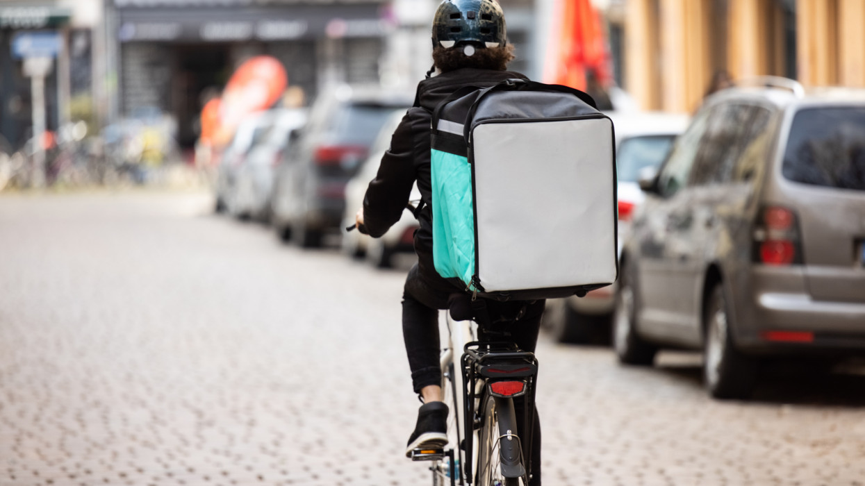 Rear view of a food delivery boy on a bike in city. Courier person riding bicycle with thermal backpack on city street for food delivery..