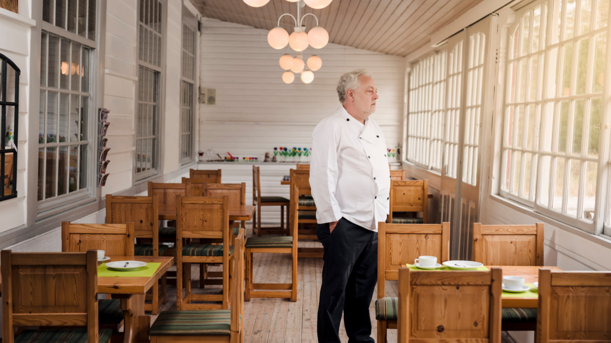 A concept to illustrate the economic impact of the Covid-19 virus on the restaurant and catering business. Restaurant owner wearing his chefâs whites standing in his empty restaurant. Photographed on location in a restaurant on the island of MÃ¸n in Denmark.