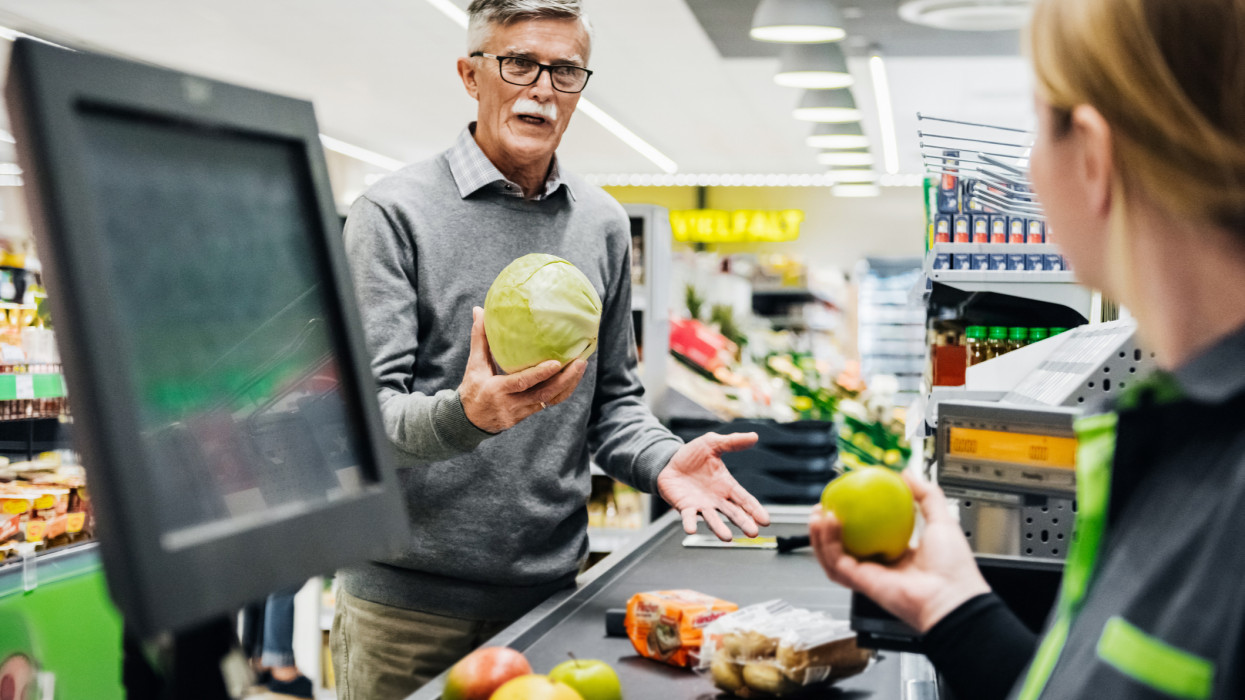 A senior man holding a melon and talking the cashier at the checkout in his local supermarket.