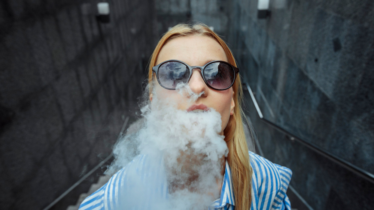 Girl in a striped stylish shirt and jeans poses on camera Day, outdoor. Blond haired girl in black glasses smoking vape