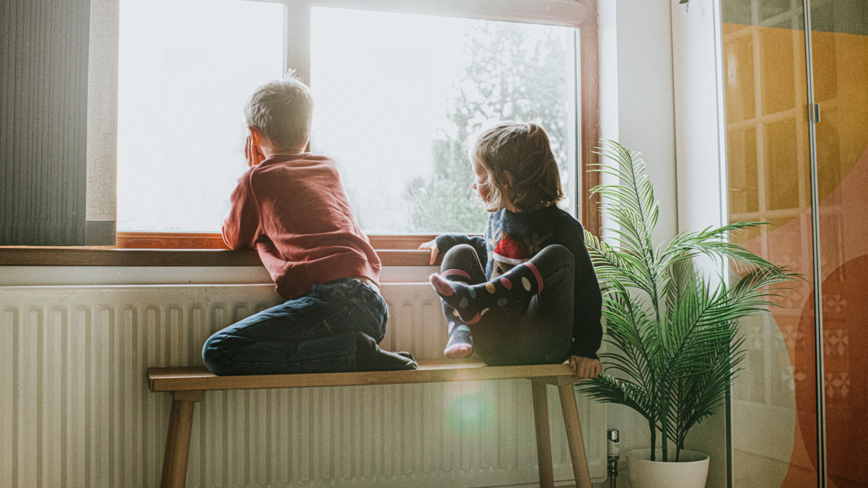 A young girl and boy sit on a wooden bench beside a long radiator and a sunny window. They gaze out through the glass and look content. Conceptual with Space for copy.