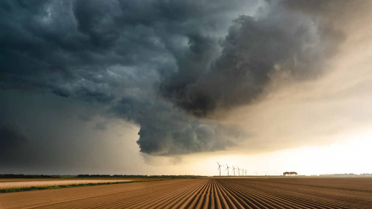 This is a picture of a storm as it is building up over an uncultivated  agricultural field and some wind turbines in the horizon. It was shot at a location in the eastern part of the Netherlands.