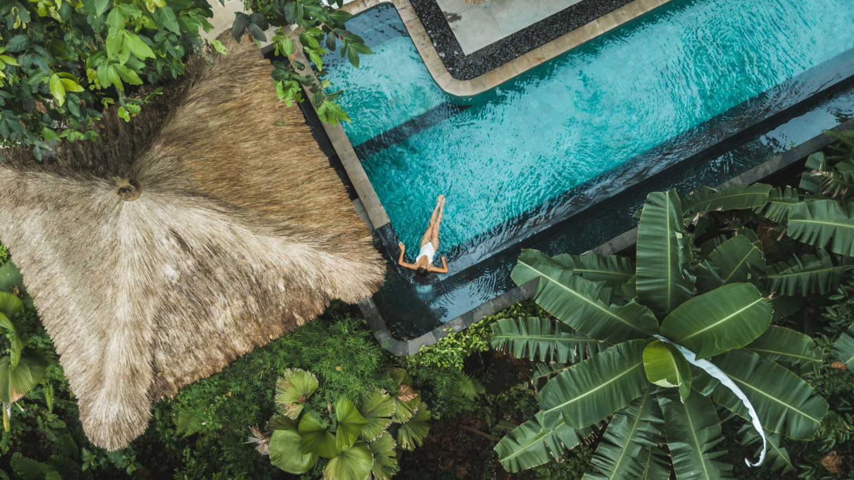 Girl relaxation in private swimming pool hiding in jungle. Wealth travel. Top view, drone point of view directly from above. Vacations in Bali. Tropical and exotic asian nature. Wellness and vitality. Balinese traditional architecture, hotel with straw roof.
