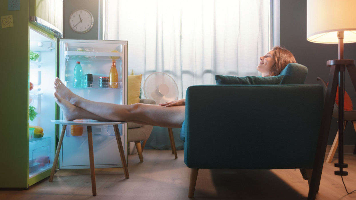 Woman cooling herself in front of the open fridge at home during the summer, she is sitting on the sofa with feet up