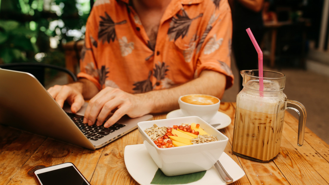 Close up image of a young freelancer, digital nomad man, sitting in a cafe in Thailand, on the Phi Phi islands, finishing up his work on the laptop while having a cup of coffee and oatmeal with fruits.
