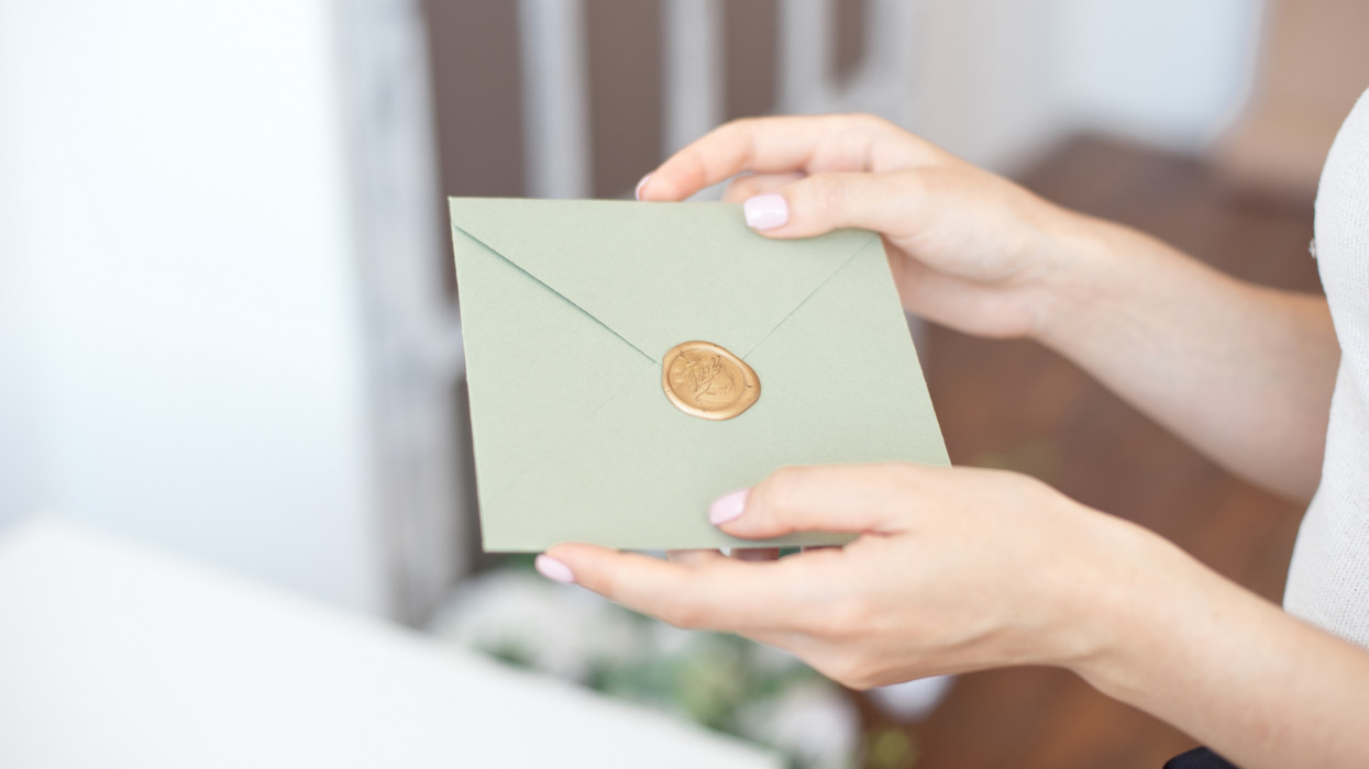 Close-up photo of female hands holding invitation envelope with a wax seal, a gift certificate, a postcard, wedding invitation card