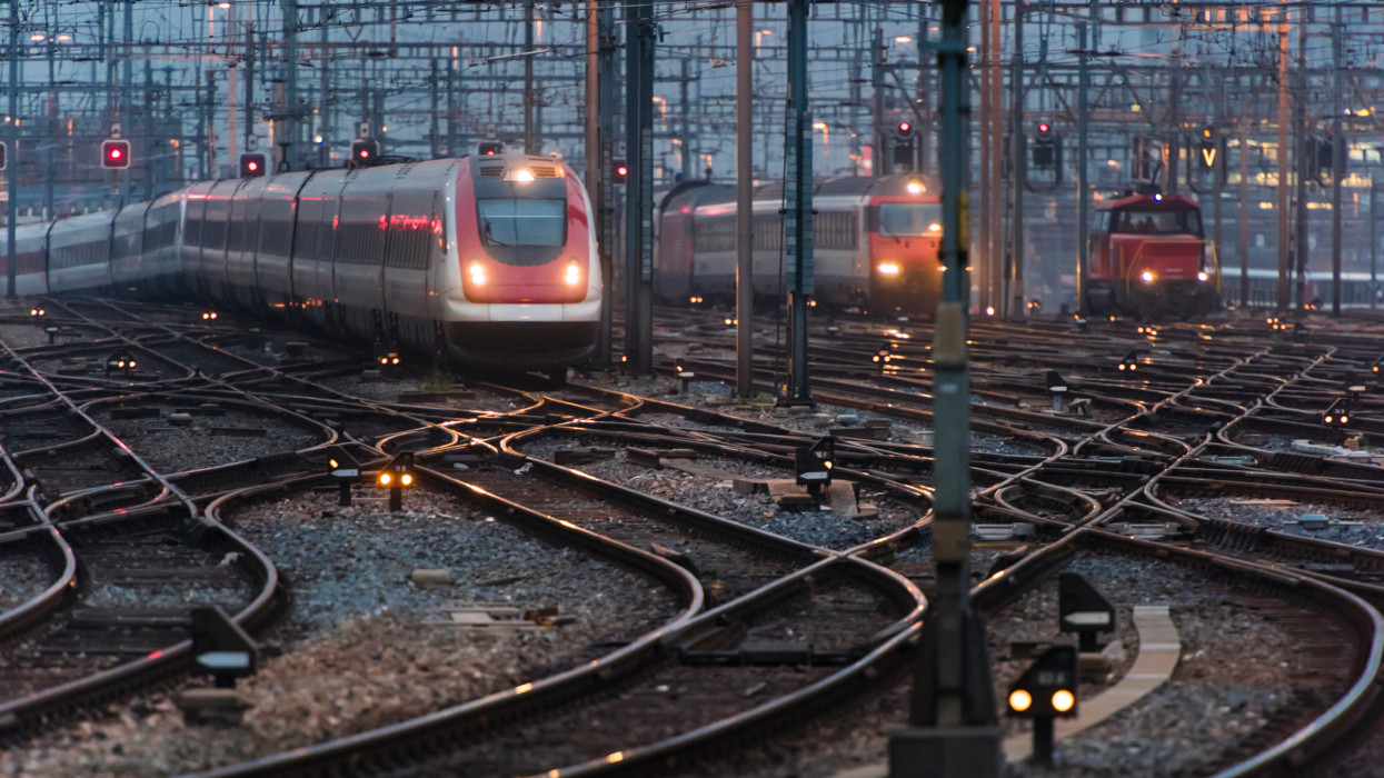In the early morning twilight, several commuter trains are approaching a busy railway station.