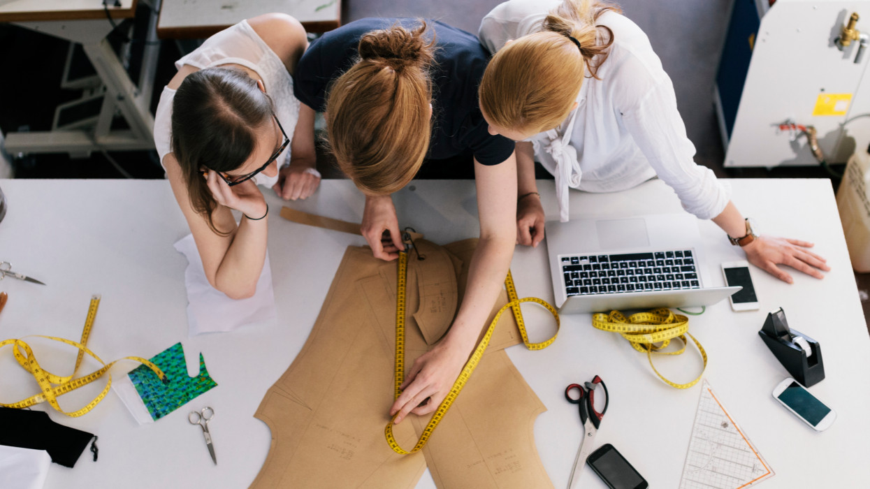 Three women fashion designers working with sewing pattern. Overhead view with tape measure, laptop, and cell phone lying on the table
