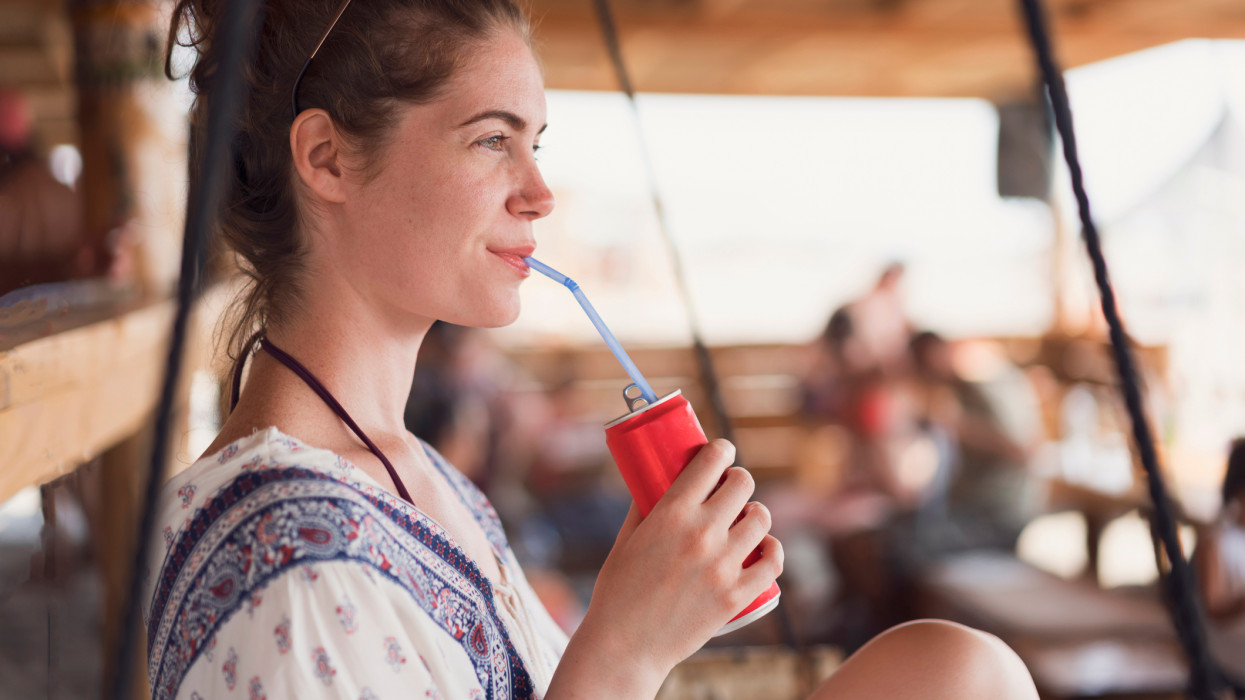 Attractive young woman relaxing on the swing at the beach bar and drinking soda.