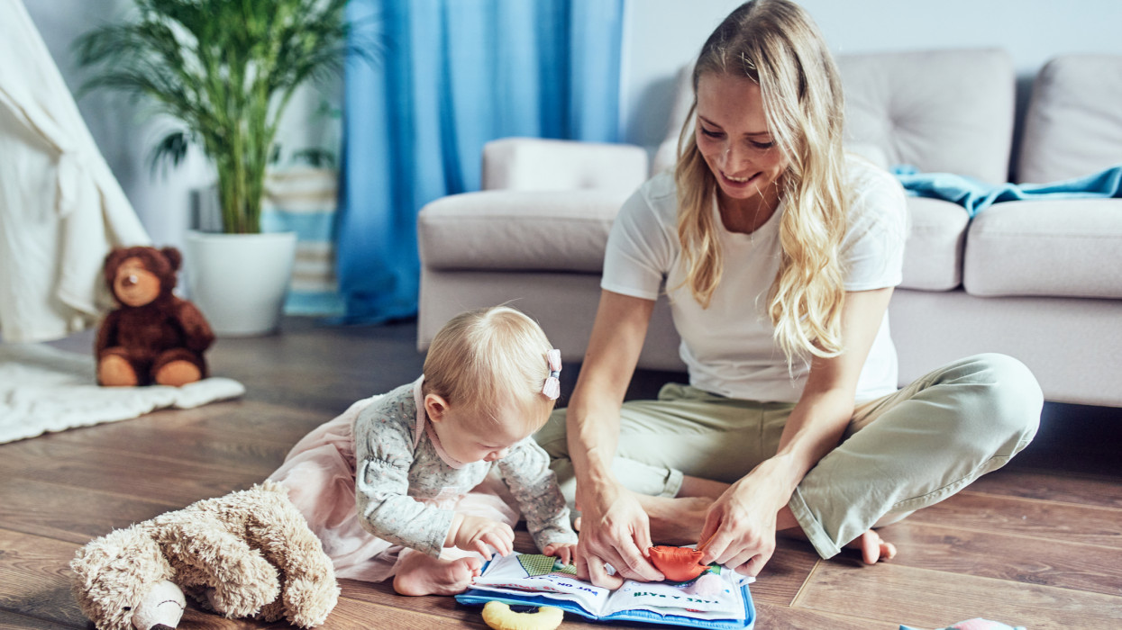A babysitter with a 1-year-old girl is reading a childrens playbook at home on the floor. A young woman with light long hair of Caucasian appearance is engaged and plays with a girl while her parents are at work
