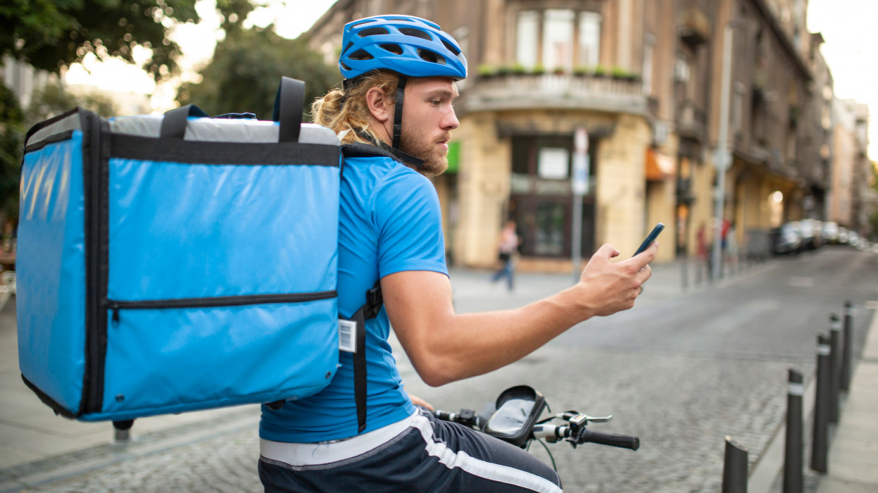 Side view of a handsome young Caucasian delivery man, wearing a blue uniform and riding a bicycle, delivering fast food around the city and using a smart phone to check for the location of his delivery