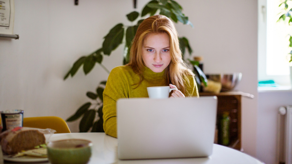 Young woman sitting at table looking at her laptop and drinking coffee. Female using laptop at home.