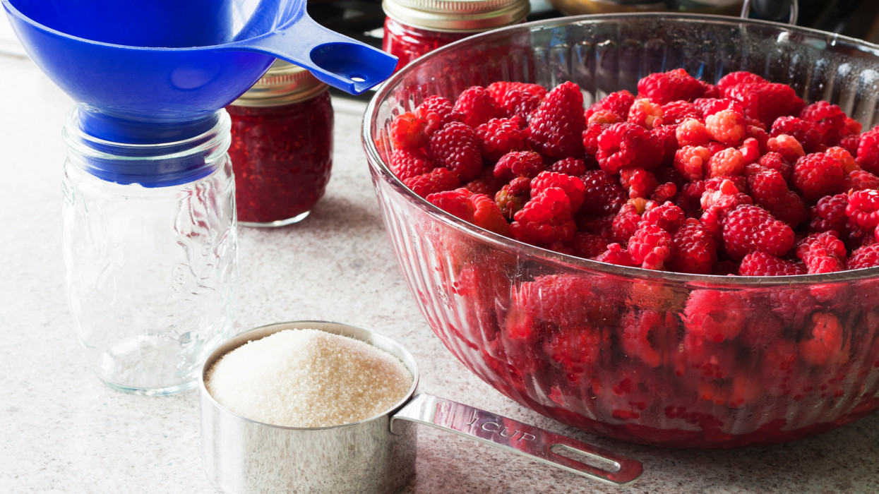 Canning raspberry jam in an average american kitchen.  A bowl full of fresh rasperries, a measuring cup full of raw sugar, an empty jelly jar with a canning funnel, completed jars and pans in the background.
