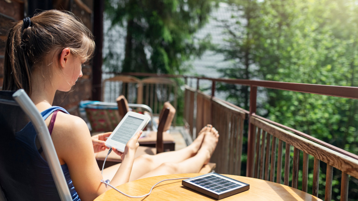 Teenage girl relaxing on balcony of a small wooden cabin in mountains. She is reading an e-book. The e-book is being charged by a small solar panel.BMPCC4K