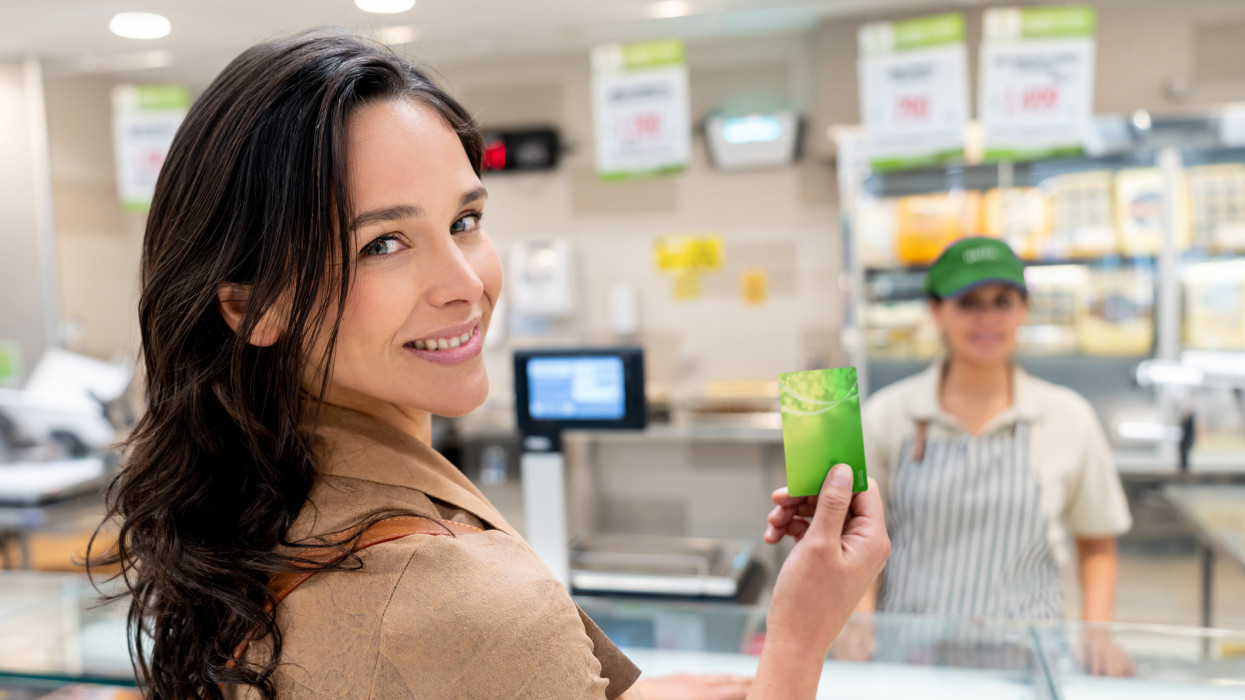 Portrait of beautiful female customer at the butchers shop holding a loyalty card smiling at camera, saleswoman standing at background shop store grocery shopping payment card paying buy