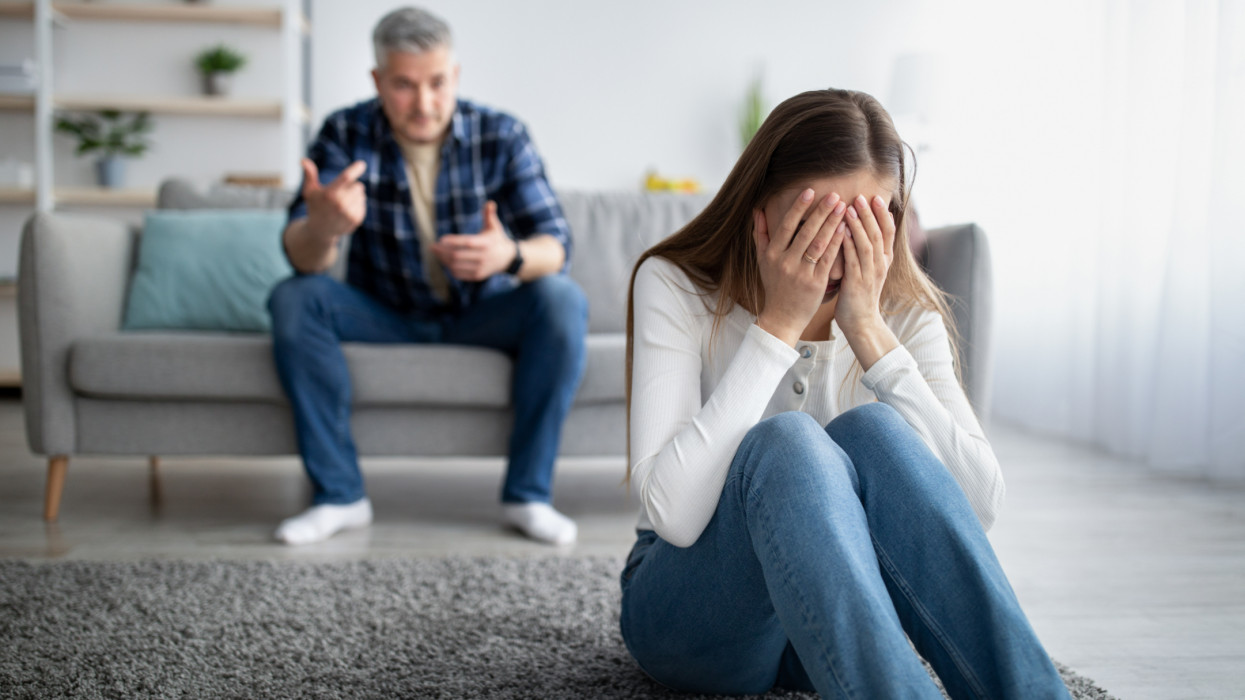 Emotional abuse in marriages concept. Mature woman crying on floor, her husband blaming or accusing her at home, copy space. Middle-aged man bullying his wife, insulting his scared depressed partner