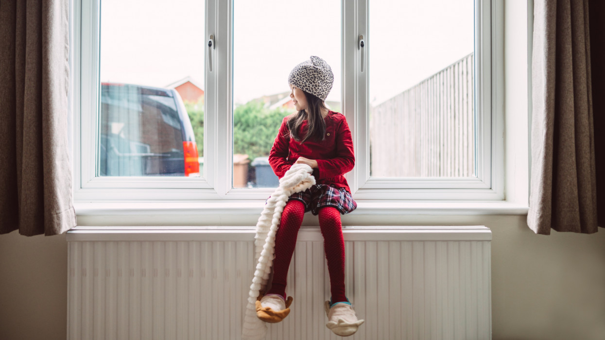 Lovely little girl in warm clothing looking through the window thoughtfully while sitting on the window sill in her new home.