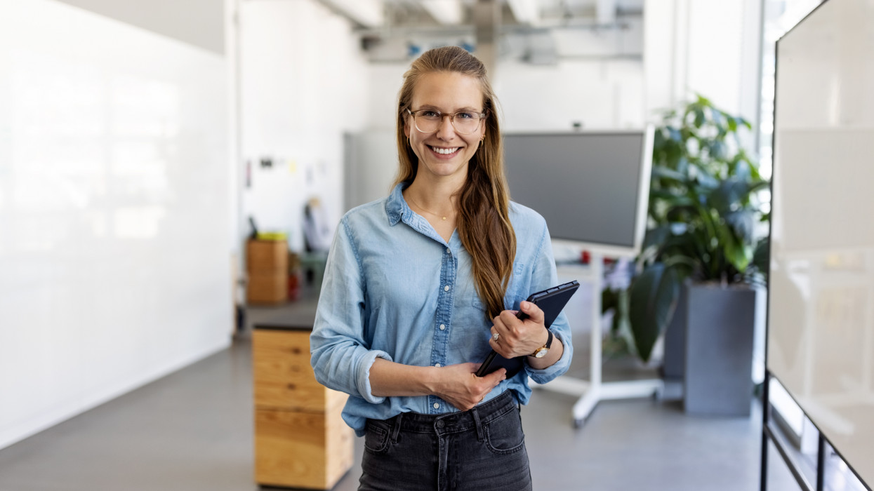 Portrait of a businesswoman with eyeglasses standing in office. Female entrepreneur looking at camera and smiling holding a digital tablet.