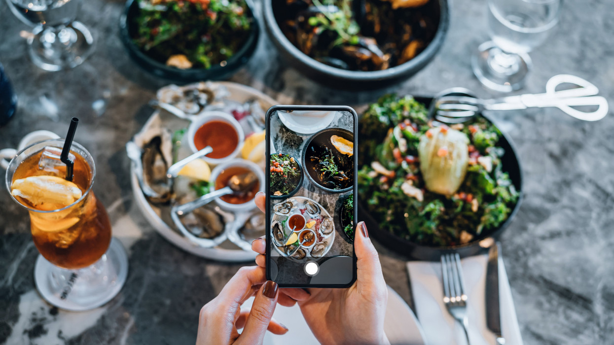 Overhead view of young woman taking photos of scrumptious and delicious meal on dining table with smartphone before eating it in restaurant. Eating out lifestyle. Camera eats first culture