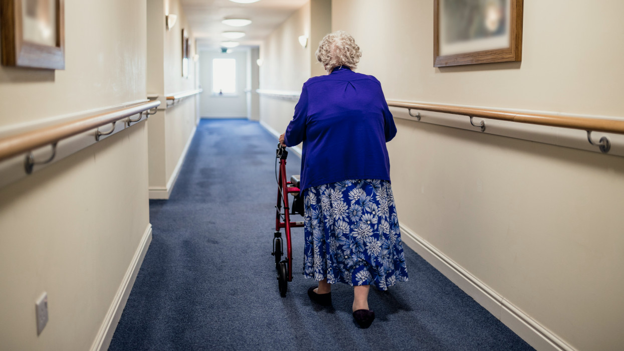A senior woman walking down a corridor with the assistance of a walker. view from rear retirement home
