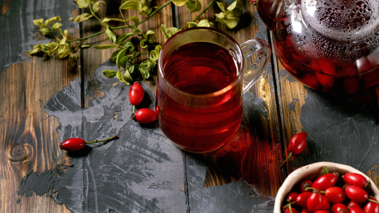 Cup of rose hip berries herbal tea and glass teapot standing on old wooden plank table with wild autumn berries around. Winter hot cozy beverage.