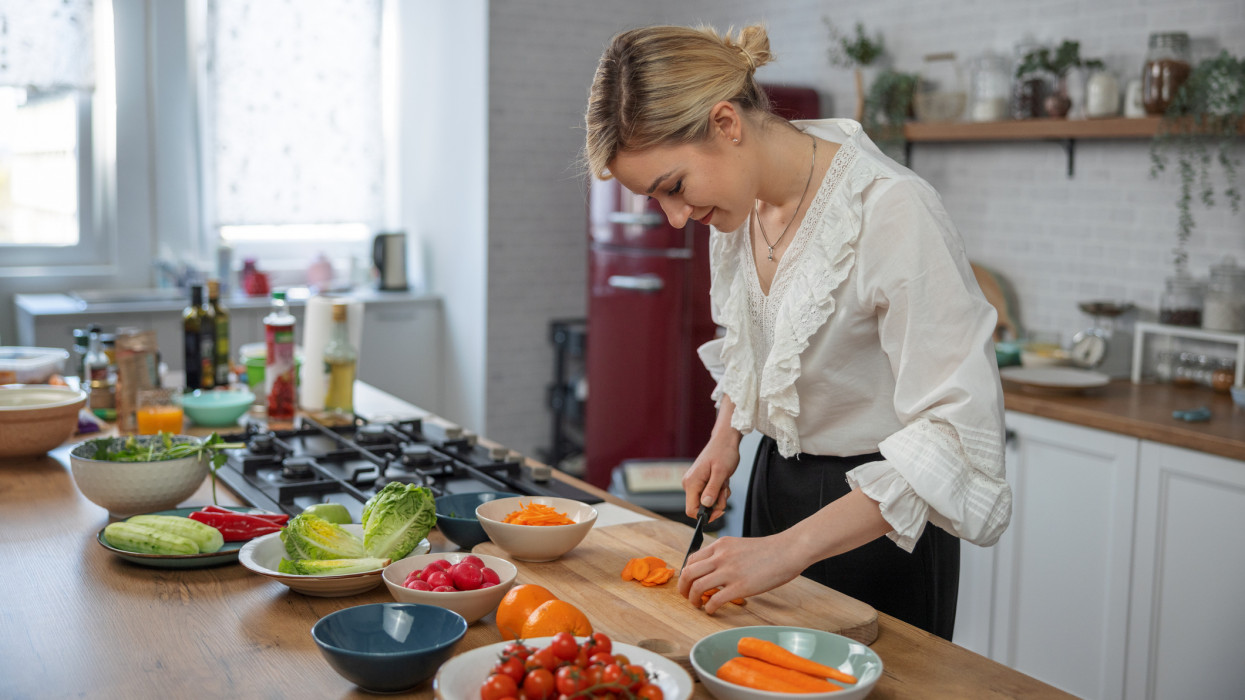 Portrait of a young caucasian woman cutting a raw carrot in the kitchen. She is smiling and preparing a salad in a wooden table in the kitchen. The table has many bowls with colorful vegetables and fruits.