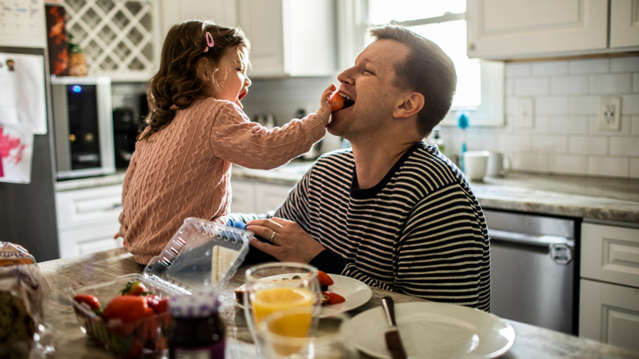 Toddler girl feeding her father a strawberry in kitchen