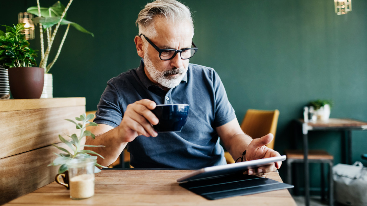 A mature man sitting in a cafe, reading on a digital tablet and drinking a cup of coffee.