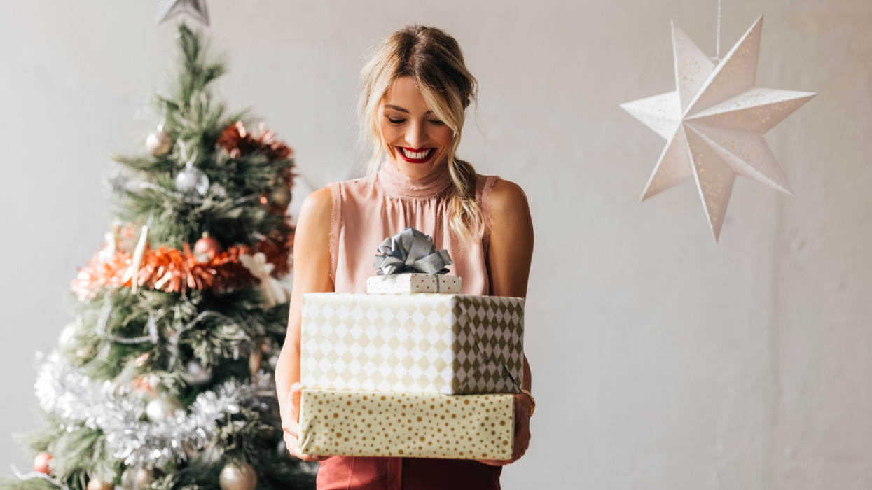 Cheerful smiling female standing next to the decorated Christmas tree and holding wrapped gift boxes in hands