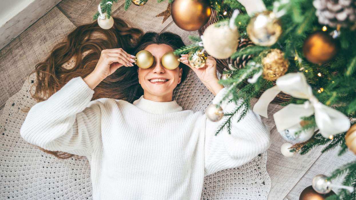 Woman in soft pastel white sweater decorating a fir tree and having fun with Christmas balls lying under the decorated branches. She is smiling happily enjoying cozy holiday time at home. December celebrations and stay home concepts