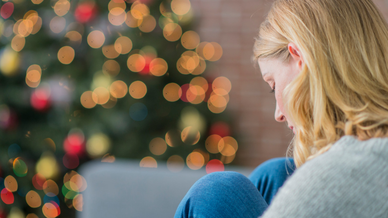 A Caucasian woman is indoors in her living room. There is a Christmas tree in the background. The woman is wearing warm clothing. She is sitting on the couch and looking sad because she is alone on Christmas day.