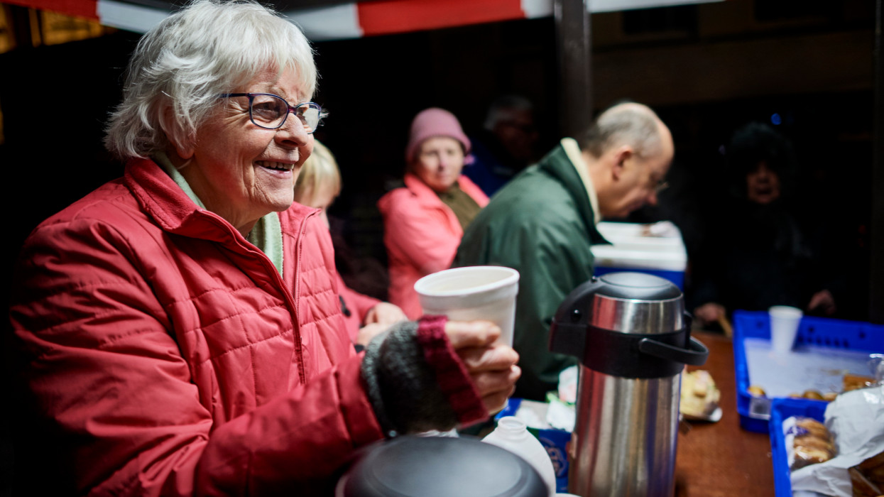 Mature female in warm clothing, serving hot drink at local voluntary soup kitchen, smiling