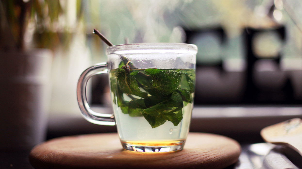 Fresh mint tea, with mint leaves and honey, in a glass cup. Stood on wooden board by a kitchen window. Plant in the background. A CAFE sign is also in background, partly covered.