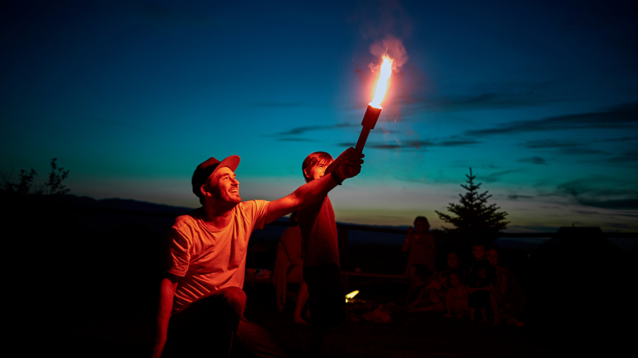 A young man holds a lit firework shooting flames into the sky while child watches