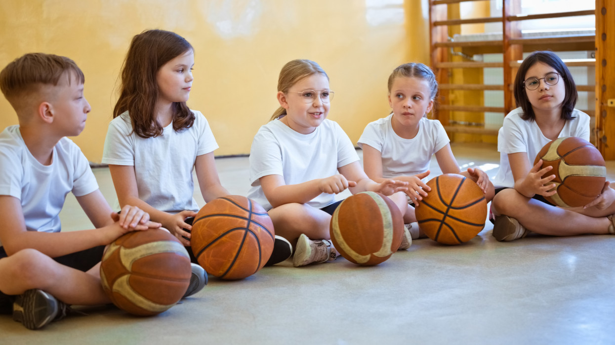 Elementary school girls and boy sitting cross legged in gym and holding basketball balls.