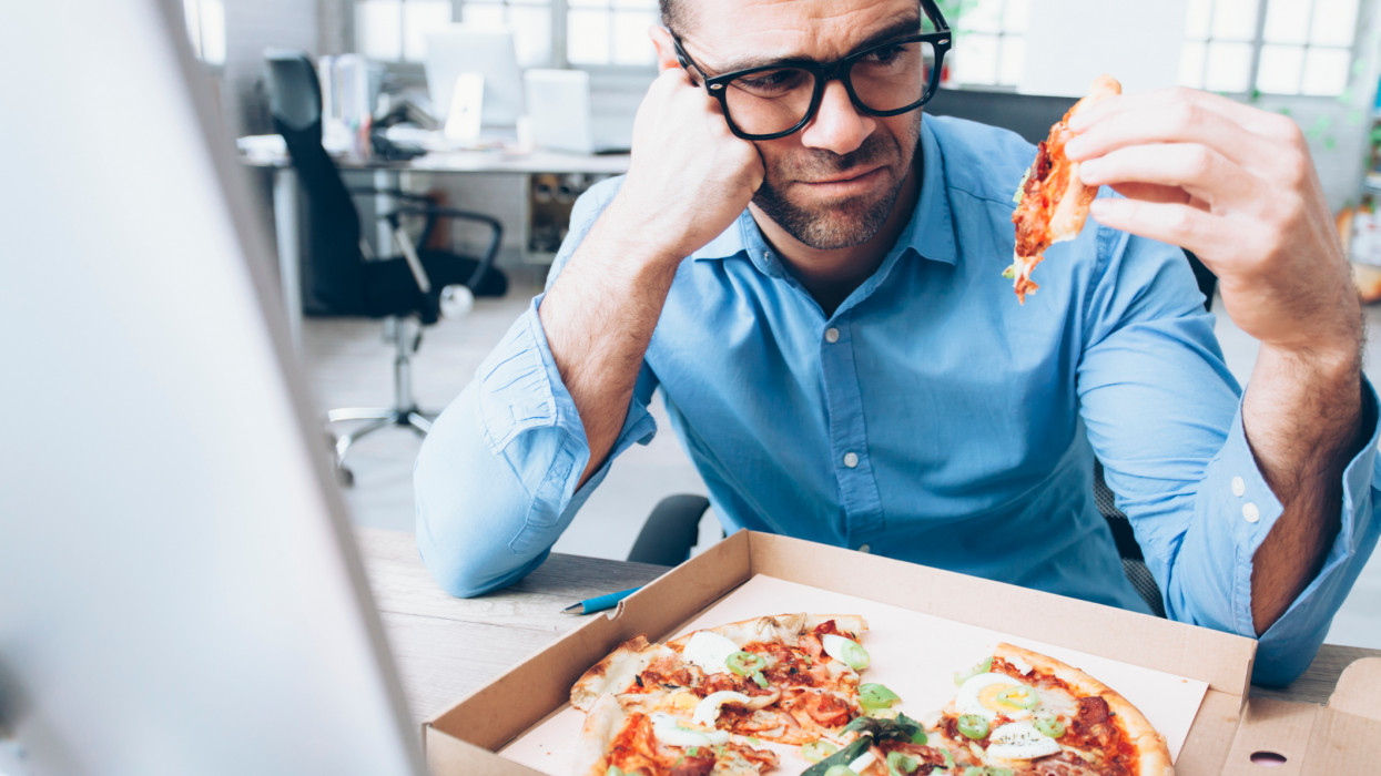 Young man with eyeglasses and shirt eating pizza at workplace. Man sitting hand on cheek, holding a slice of pizza in hand and looking at it. Pizza box on table. Desk with computers and windows on background.