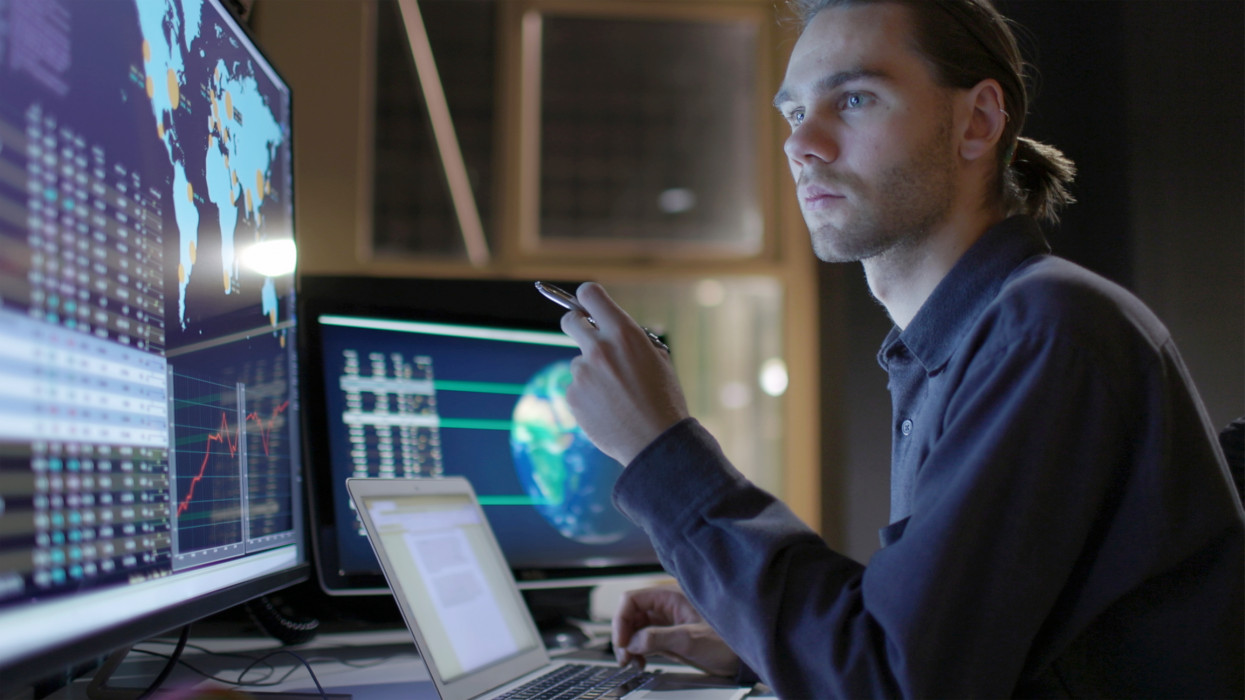 Stock photograph of a young man working with a large computer monitor, Heâs studying global data with maps of the Earth.