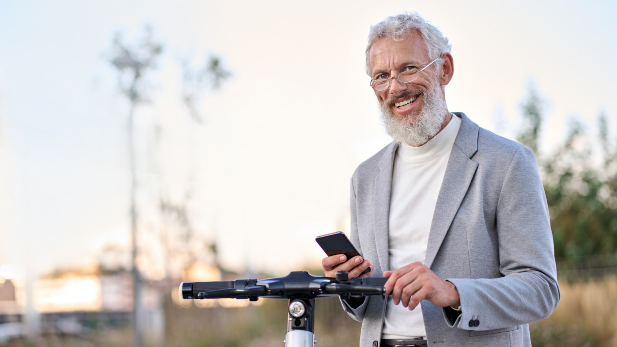 Happy old senior adult business man holding smartphone using electric bike rental digital phone mobile app renting scooter in city public eco transport mobile application standing in urban park.