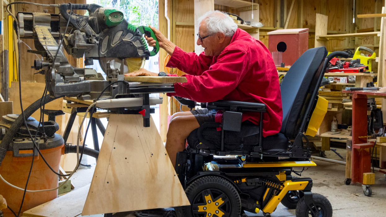 Disabled man in wheelchair operates a table saw, cutting a piece of wood, in his home workshop.
