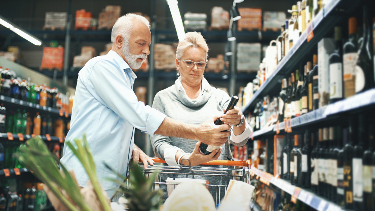 Closeup side view of early 60s couple walking through supermarket aisle and choosing some wine for tonights dinner. They are holding a bottle of rose wine and reading the label. Shelves stacked with wines are right next to them, brand free, a bit blurred.
