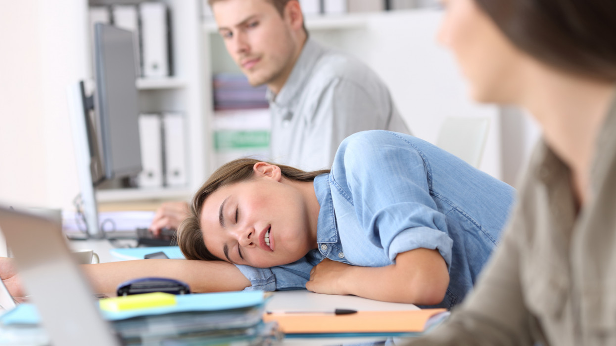 Fatigued employee sleeping on a desktop at office with her colleagues looking