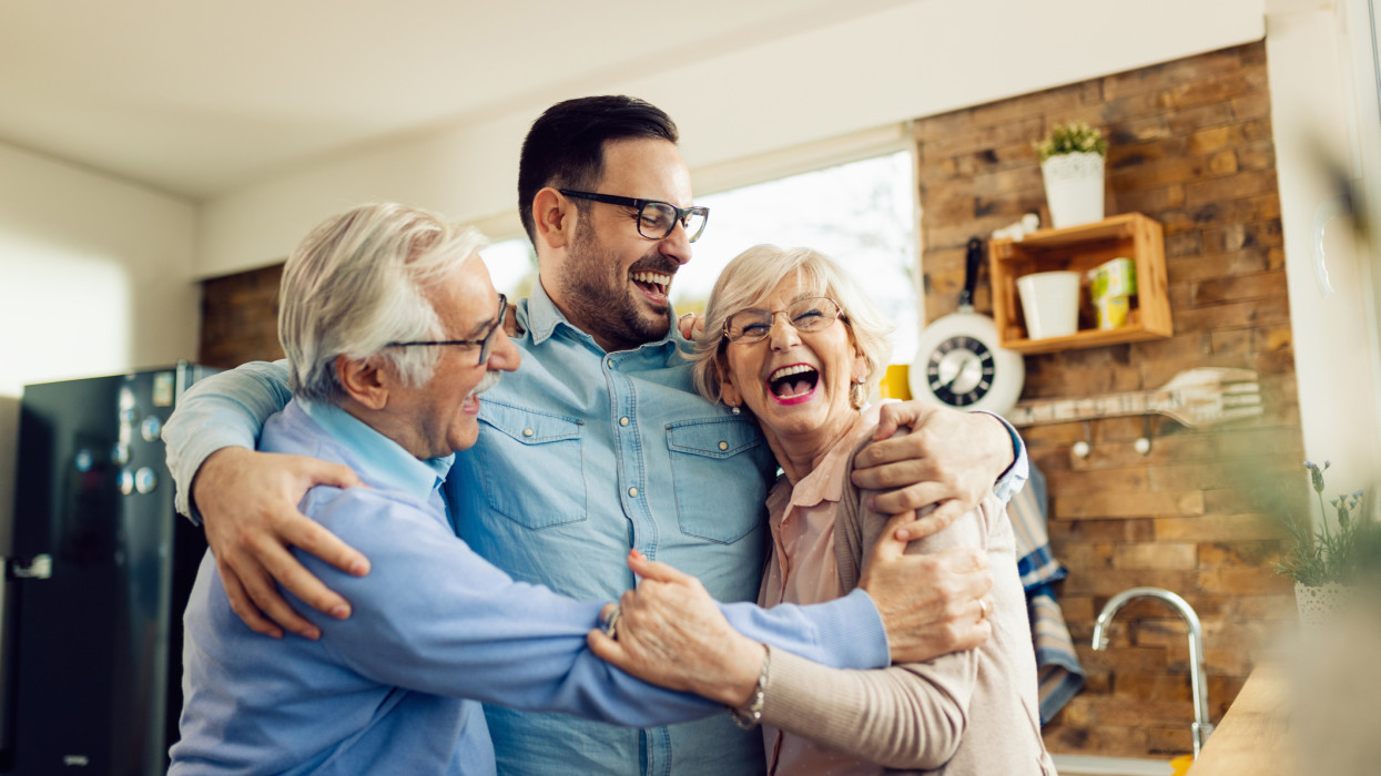 Cheerful mid adult man and his senior parents laughing while embracing in the kitchen.