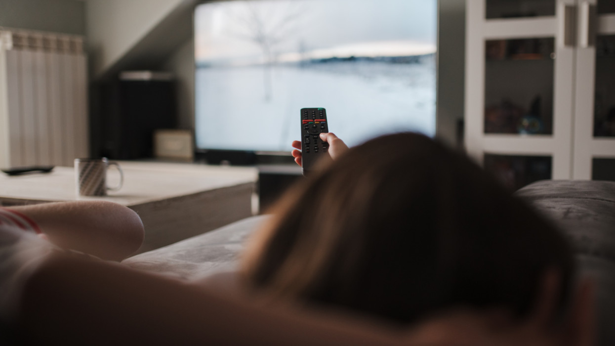 View from behind of a young Caucasian girl lying on the sofa holding the TV remote control. She is relaxing on the sofa at home. Unrecognizable girl.