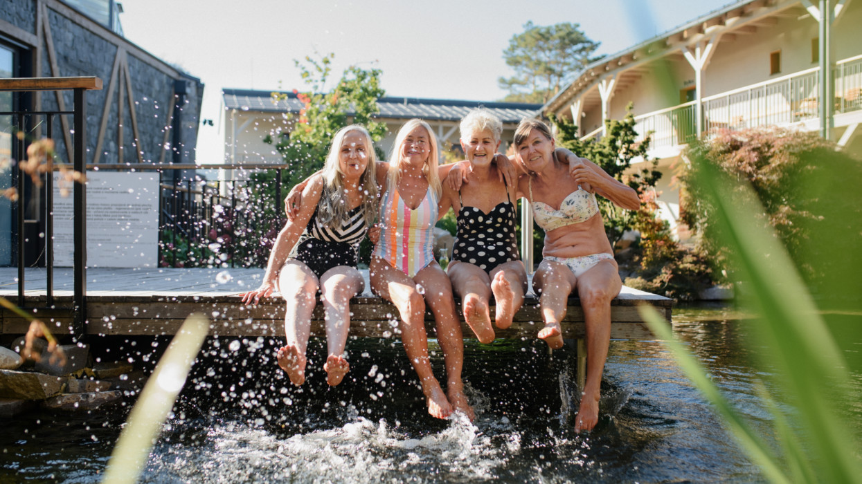 Happy senior women friends in swimwear sitting on jetty and splashing water outdoors by swimming pool.