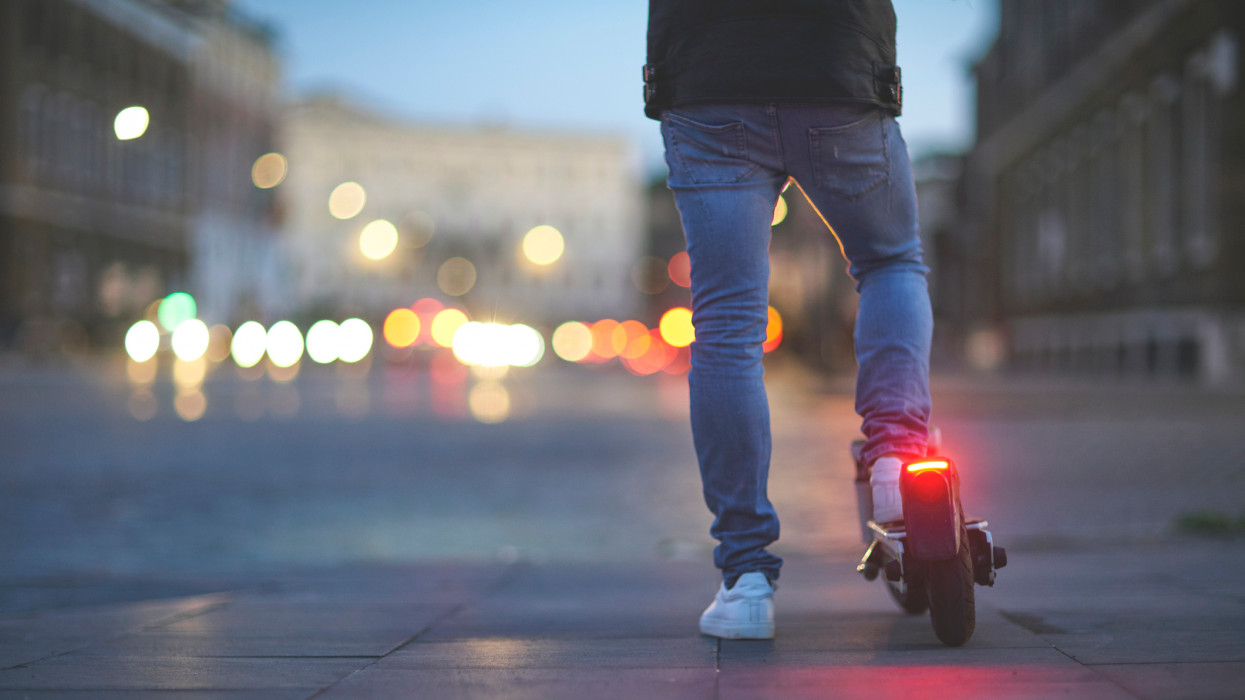 Low section of man riding electric push scooter. Traffic street lights on background.