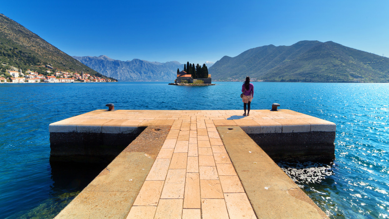 Lone female tourist standing on dock at Our Lady of the Rocks (Gospa od Skrpjela) man-made island, looking across water to Sveti Dorde Island with coastal town of Perast in distance on Bay of Kotor, Montenegro.