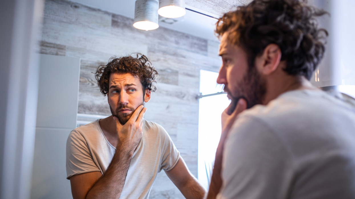 Morning hygiene, Handsome man in the bathroom looking in mirror. Reflection of handsome man with beard looking at mirror and touching face in bathroom grooming