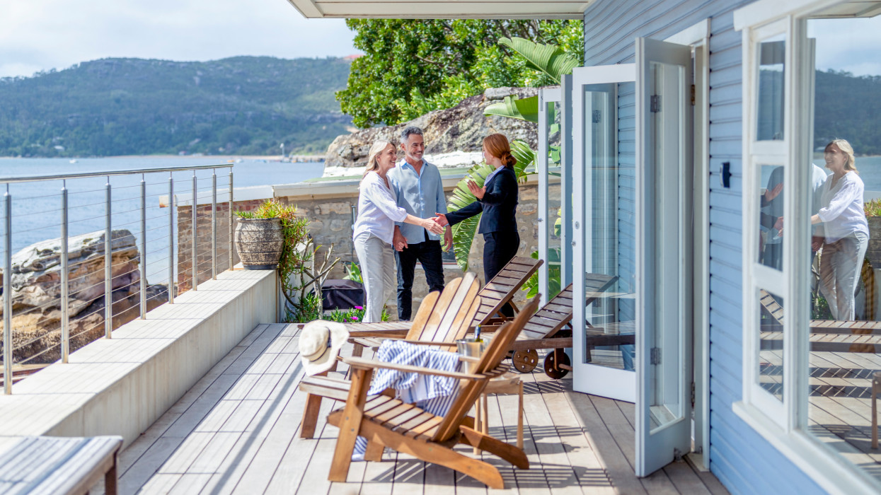 Real estate agent showing a mature couple a new house. The house is contemporary. All are happy and smiling and shaking hands. The couple are casually dressed and the agent is in a suit. Waterfront can be seen in the background