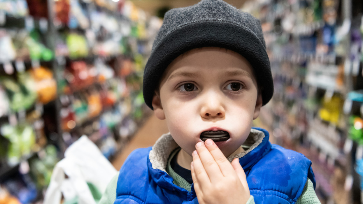 Caucasian three years old little boy sitting in a shopping cart at a supermarket aisle having a chocolate cookie wearing a toque hat
